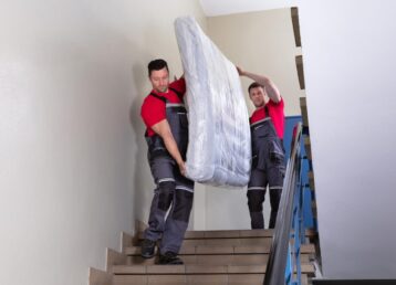 Two,Young,Male,Movers,In,Uniform,Carrying,The,Wrapped,Mattress